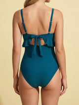 Back view of a woman wearing a teal one-piece swimsuit with a tie-back closure and ruffle trim.