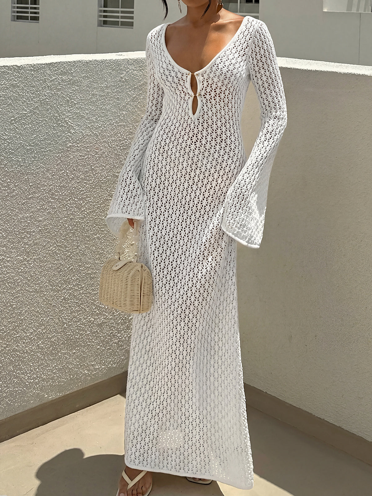 Woman wearing a white open knit maxi dress with bell sleeves and a deep V-neckline, holding a straw handbag.