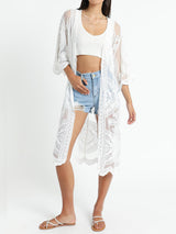 Woman wearing a white lace kimono over a white crop top and denim shorts, with white sandals.