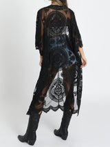Back view of a woman wearing a black lace kimono over a white crop top and denim shorts, with black cowboy boots.