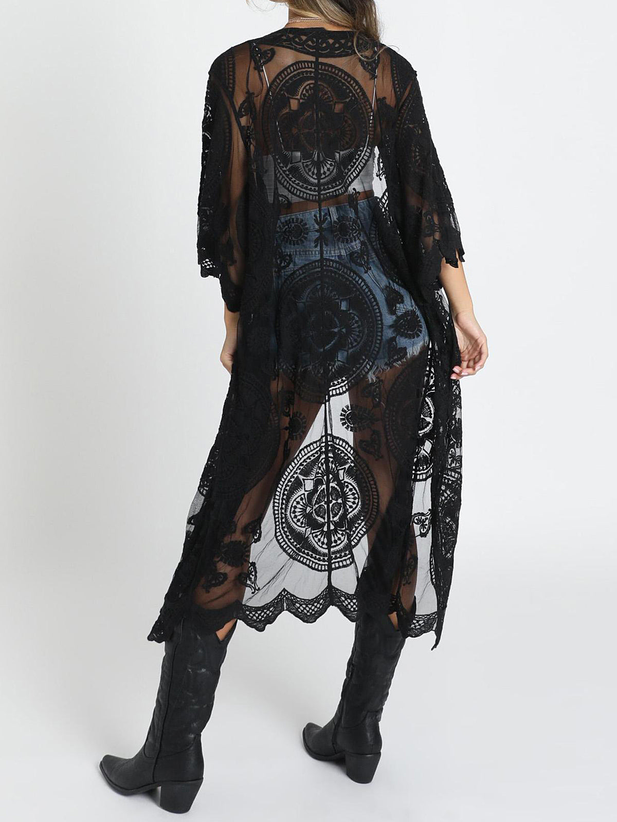 Back view of a woman wearing a black lace kimono over a white crop top and denim shorts, with black cowboy boots.