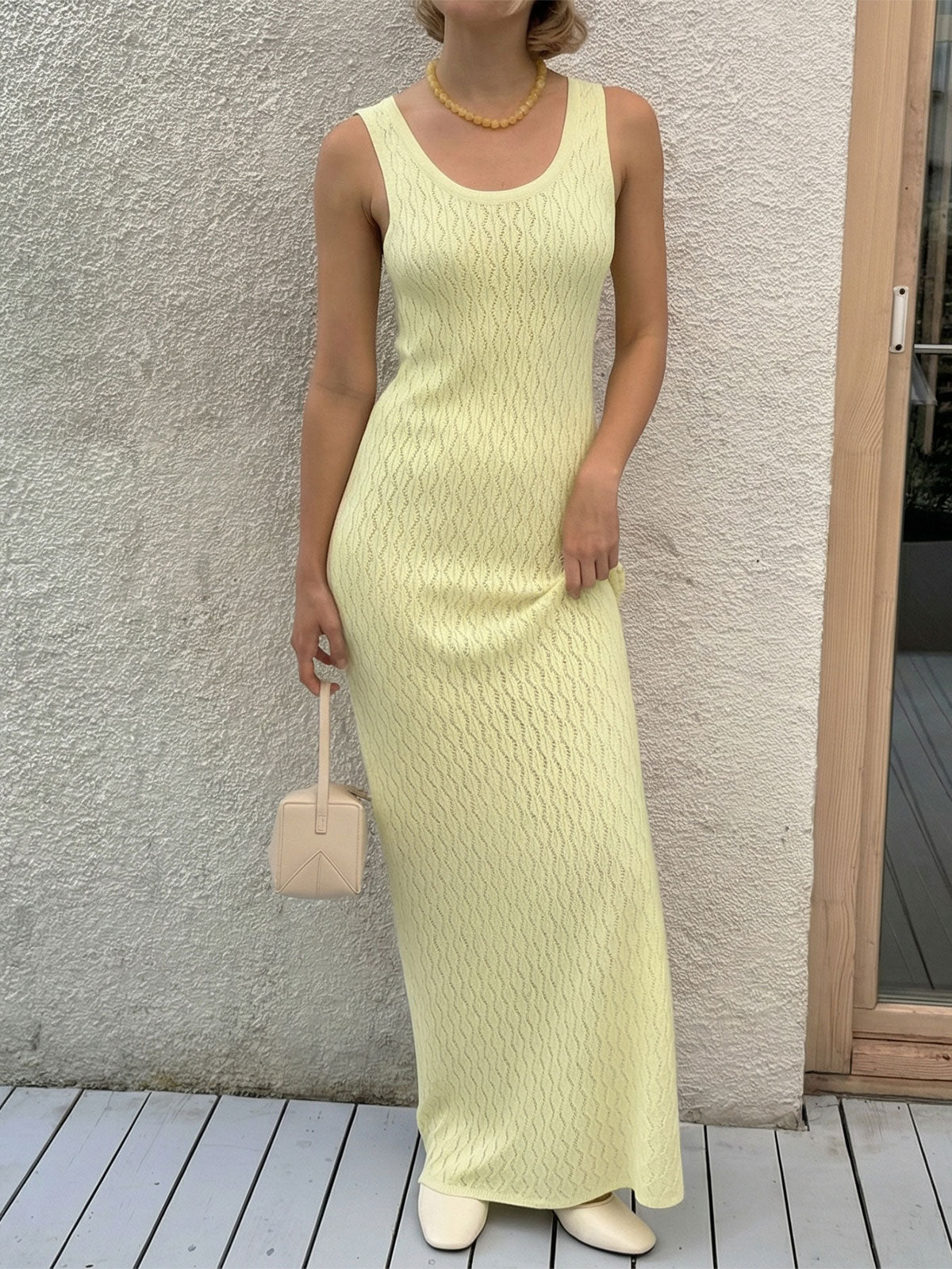 Woman wearing a pale yellow sleeveless maxi knit dress with a scoop neck, holding a cream-colored mini handbag.