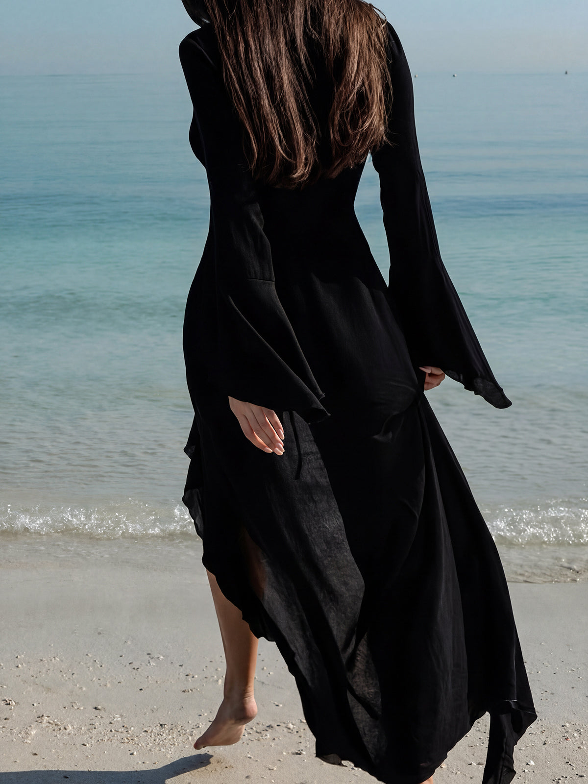woman from behind in a flowing black maxi dress with ruffled trim and bell sleeves walking on a beach