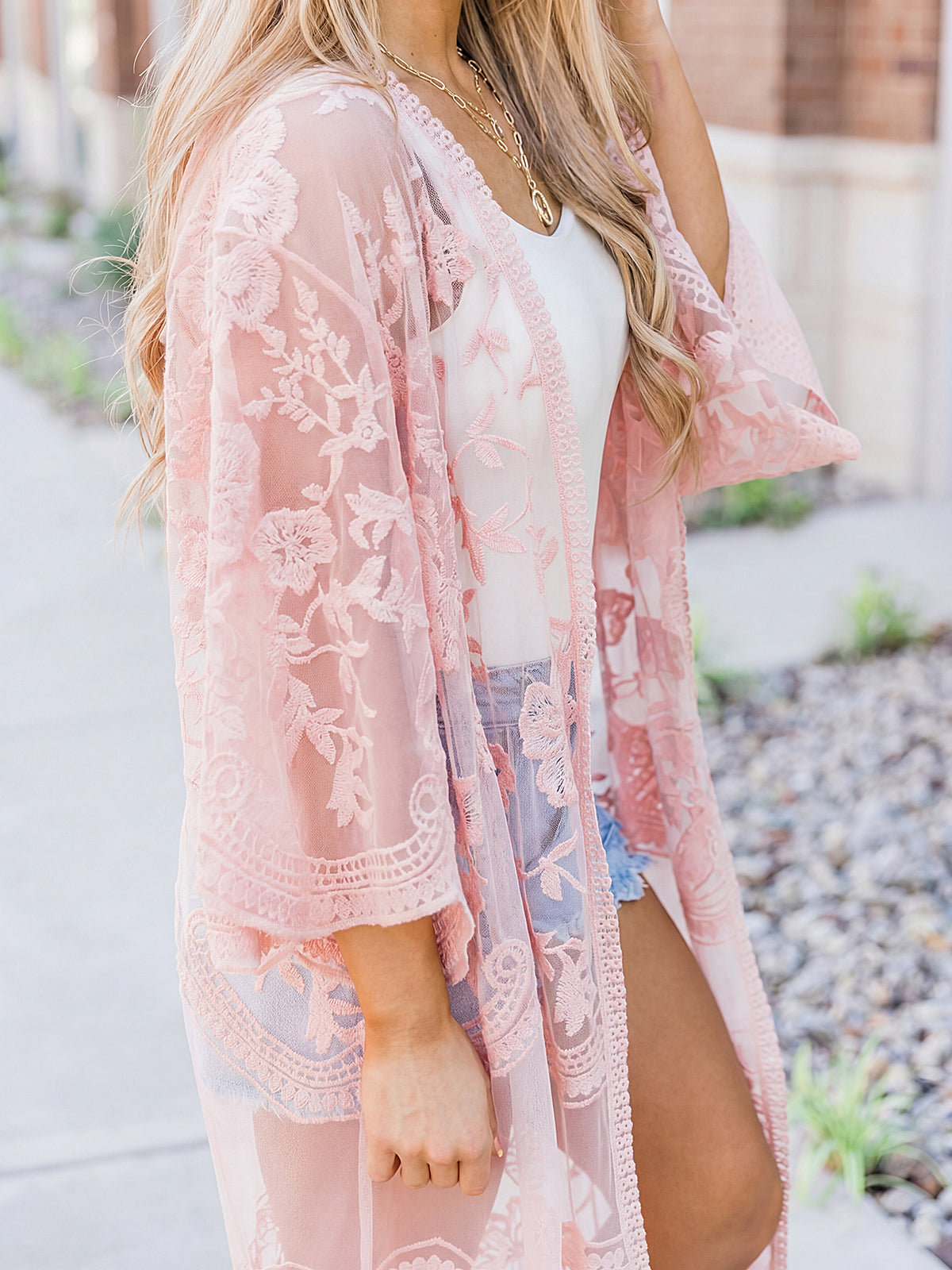 Woman wearing a pink lace kimono over a white top and denim shorts.