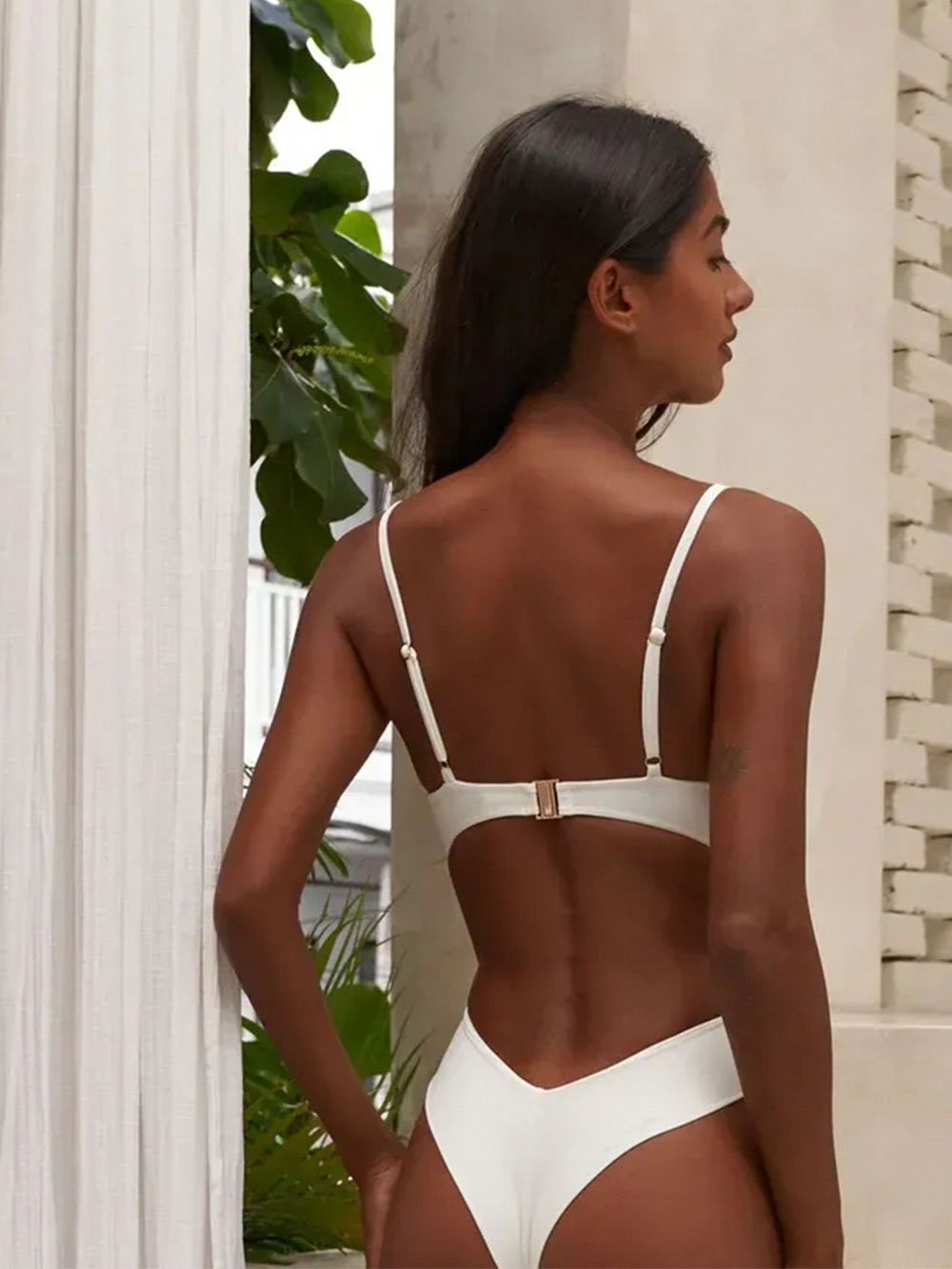 Back view of a woman wearing a cream one-piece swimsuit with adjustable straps and gold hardware