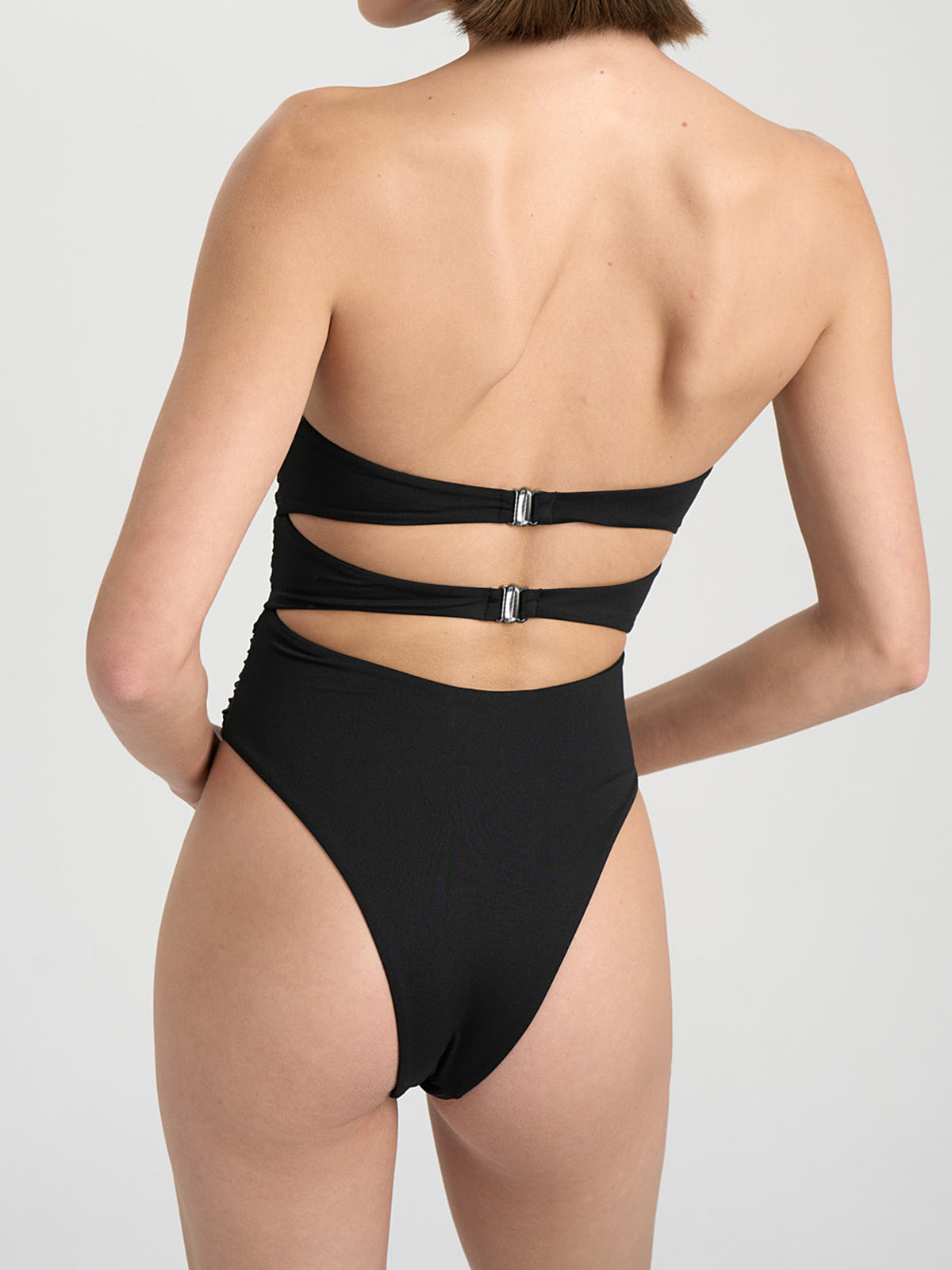 Woman wearing a black strapless one-piece swimsuit with cut-out details and floral embellishments, side view.