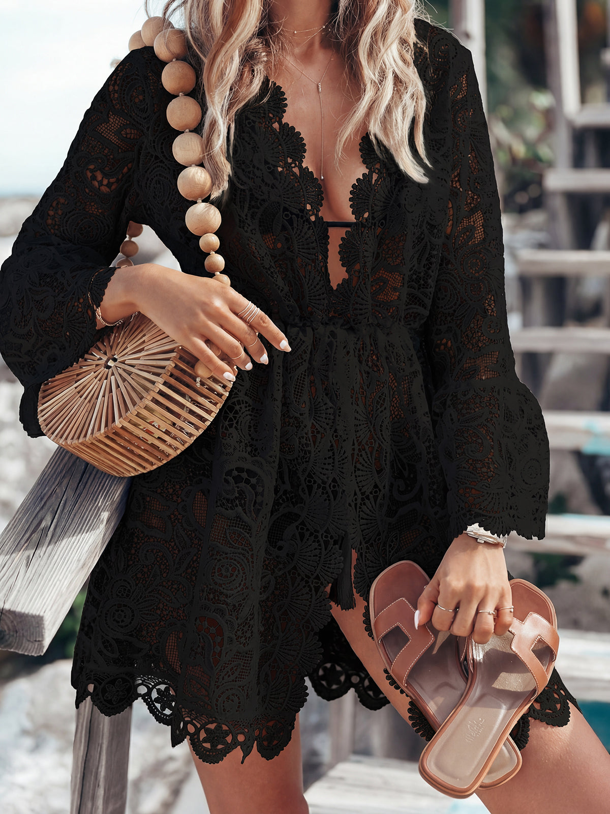 Woman wearing a black lace tunic over a swimsuit, holding a woven bag and sandals.